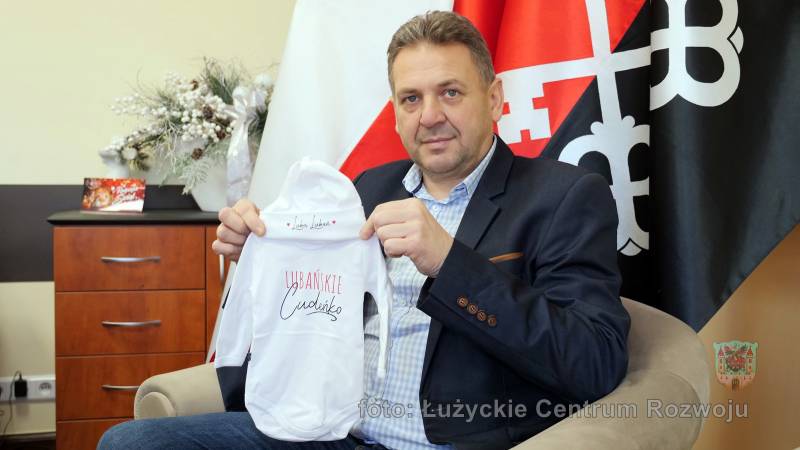 burmistrz Lubania siedzi na tle flagi Polski i flagi Miasta Lubań, w rękach trzyma body i czapeczkę dla niemowlaka, na czapeczce napis "Lubię Lubań", a na body "Lubańskie Cudeńko"