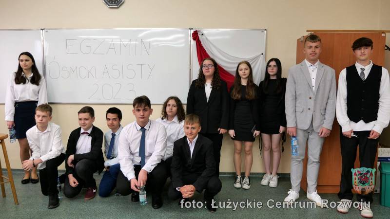 grupa uczniów i uczennic, za nimi tablica z napisem "egzamin ósmoklasisty 2023"